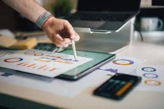 A hand uses a tablet beside printed financial charts at a desk.