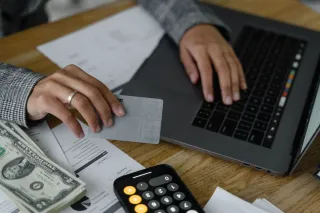 A person holds a bank card beside a laptop, dollar bills, printed paperwork and a phone calculator.