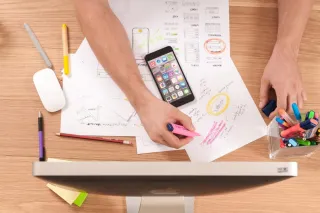 Hands mark interface sketches with a highlighter beside a phone and computer on a desk.