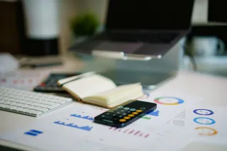 Printed bar and doughnut charts spread across a desk beside a phone calculator, open notebook and computer keyboard.