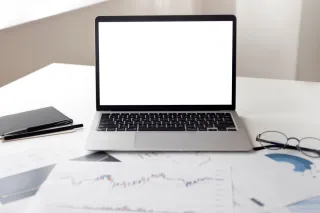 A laptop with a blank screen sits among printed financial line charts and allocation-style circular charts on a white desk.