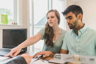 A couple reviews documents with another person across a desk, with a calculator and laptop nearby.