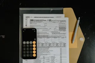 A smartphone calculator and gray pen resting on blank US income-tax forms and a yellow folder on a dark desk.
