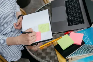 A person writes a note in an open notebook beside a laptop, colored sticky notes, pens and pencils.