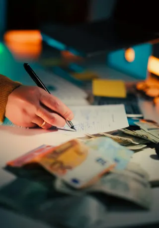A hand writing on a document beside euro and US dollar banknotes, with a laptop in the background.