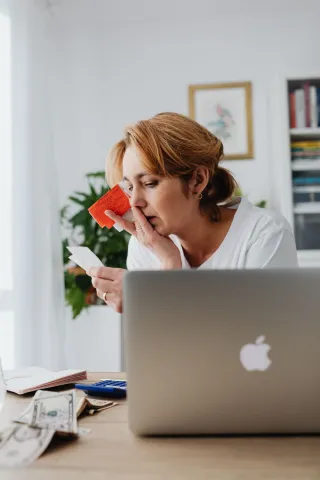 A woman holds receipts and a small card while reviewing paperwork beside a laptop and calculator at home.