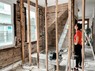 A woman inspects a room with exposed wooden studs, stripped walls, a ladder and renovation debris.