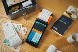 A payment terminal prints a receipt beside a cash box, Czech banknotes and paper receipts on a wooden table.