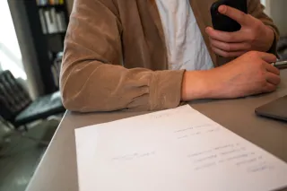 A person holds a phone beside paperwork and a laptop on a desk.