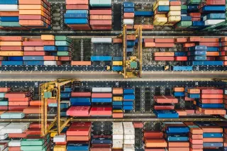 Overhead view of multicolored shipping containers stacked in rows around cargo cranes and marked port lanes.